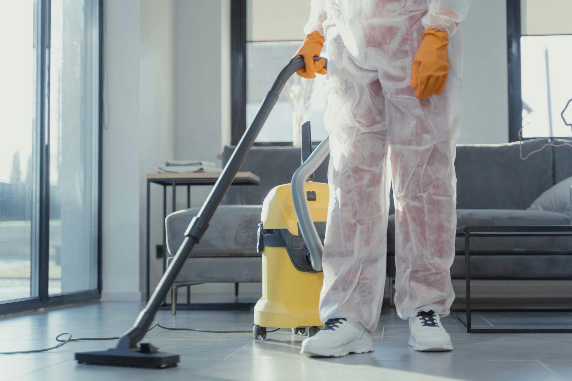 Un agent d'entretien, vêtu d'une combinaison de protection et de gants, passe l'aspirateur dans un salon moderne.